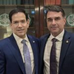 Dois homens de terno posam juntos em frente a uma estante com objetos decorativos
