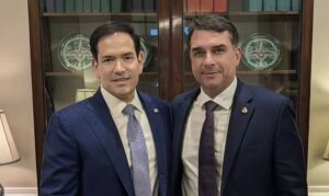 Dois homens de terno posam juntos em frente a uma estante com objetos decorativos