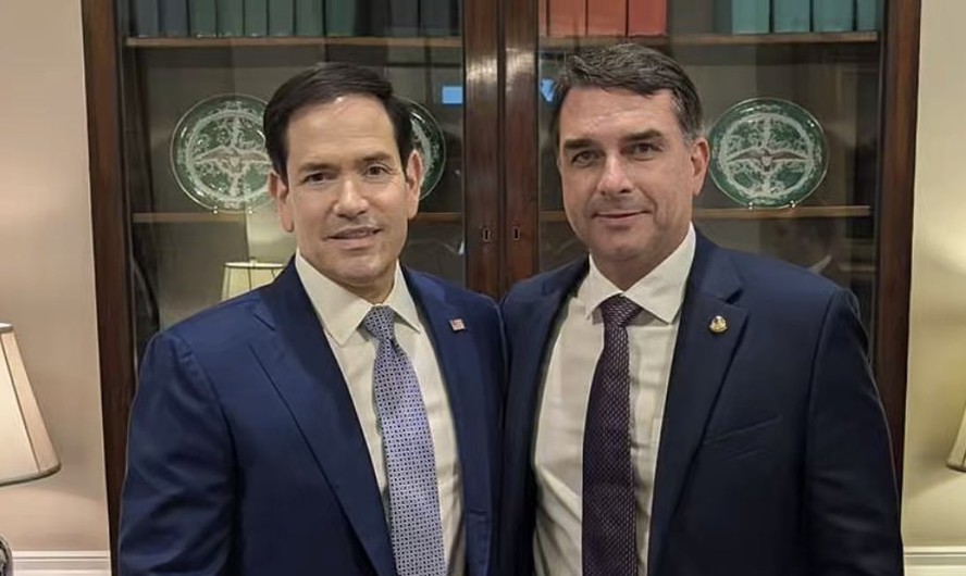 Dois homens de terno posam juntos em frente a uma estante com objetos decorativos