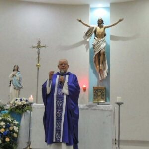 Um homem em vestes religiosas fala em frente a um altar com imagem de Cristo e estátua de Nossa Senhora