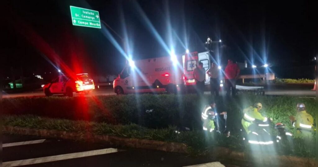 Um grupo de pessoas atende uma ocorrência na beira de uma rodovia à noite com viaturas e uma moto derrubada