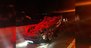 Carro capotado de lado na pista à noite com luzes de emergência e equipamento de resgate ao fundo
