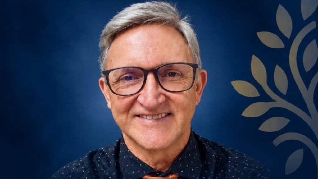 Um homem de óculos e camisa escura posa em frente a fundo azul com desenho de folhas