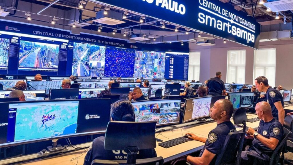 Um grupo de pessoas trabalha em uma central de monitoramento com telas exibindo mapas e imagens de câmeras