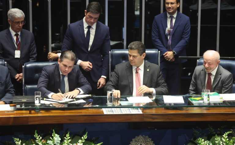 Um homem assina documento em mesa com outros homens em fundo formal