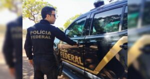 Um homem com uniforme da Polícia Federal abre a porta de uma viatura em área externa com árvores ao fundo