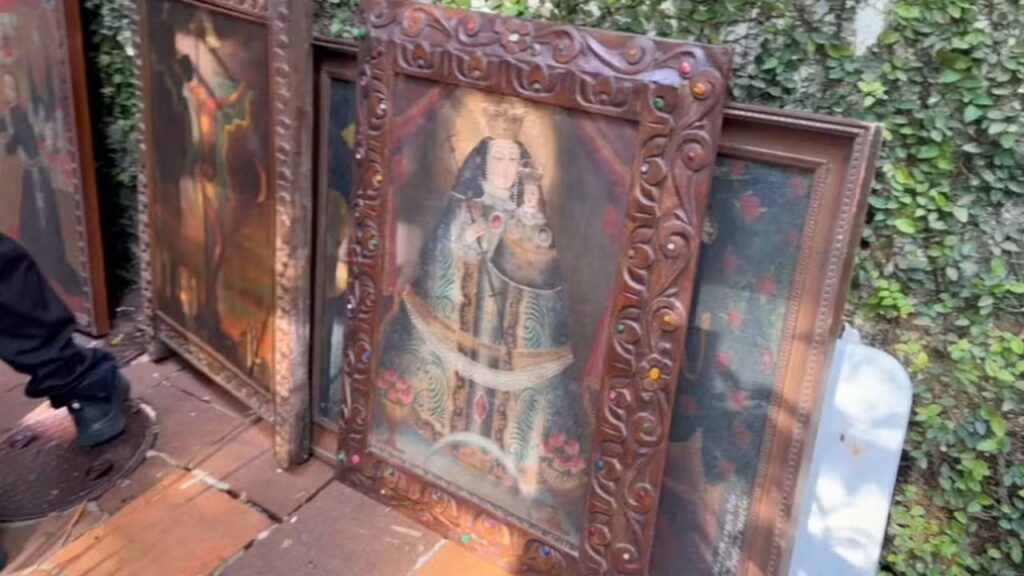 Várias pinturas religiosas em molduras ornamentadas apoiadas no chão ao lado de uma parede com vegetação
