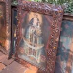 Várias pinturas religiosas em molduras ornamentadas apoiadas no chão ao lado de uma parede com vegetação
