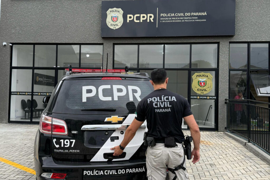 Um homem de uniforme se aproxima de uma viatura da Polícia Civil em frente a um prédio da corporação