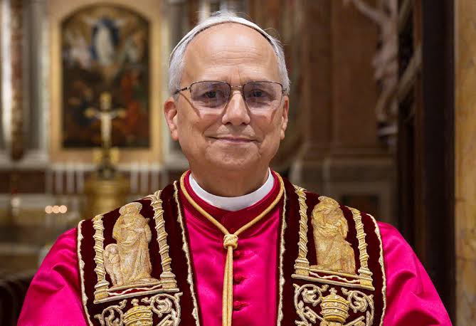 Um homem de vestes religiosas ornamentadas posa em frente a um altar com pintura e cruz ao fundo