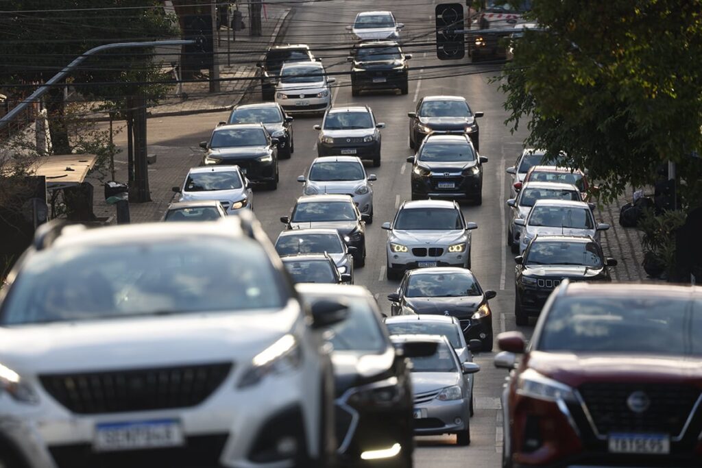Trânsito intenso com diversos veículos em movimento lento em via urbana com semáforos e árvores ao lado