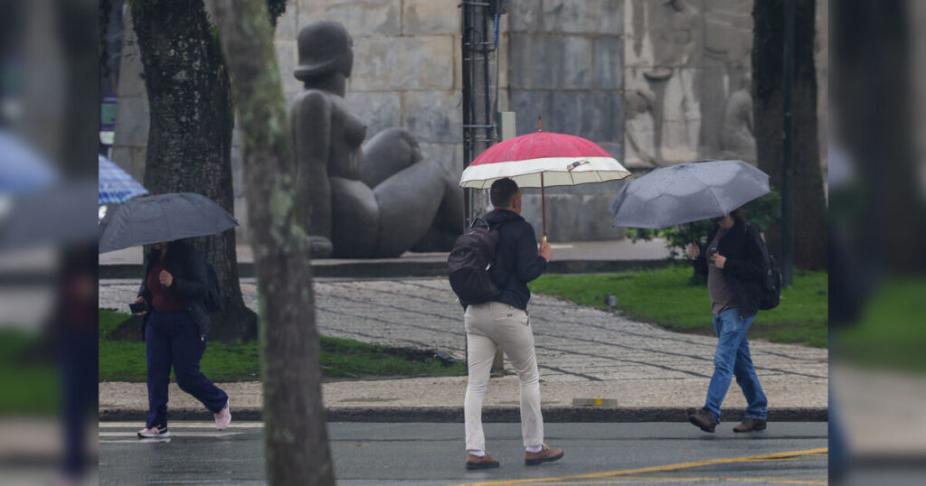Pessoas caminham com guarda-chuvas em calçada molhada perto de estátua ao fundo