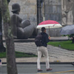 Pessoas caminham com guarda-chuvas em calçada molhada perto de estátua ao fundo
