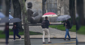 Pessoas caminham com guarda-chuvas em calçada molhada perto de estátua ao fundo