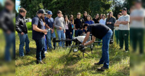 Um grupo de pessoas observa um homem ajustando um drone em um campo verde