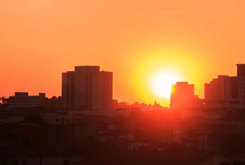 Pôr do sol sobre silhuetas de prédios em meio a céu alaranjado e intenso calor