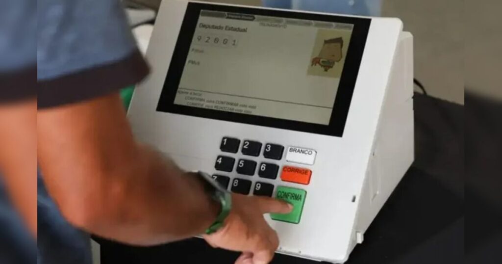 Mão apertando botão verde de confirmação em urna eletrônica com número de candidato exibido na tela