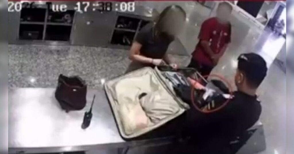 Um homem de terno revista uma mala aberta em uma mesa com duas pessoas ao fundo