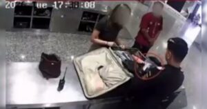 Um homem de terno revista uma mala aberta em uma mesa com duas pessoas ao fundo
