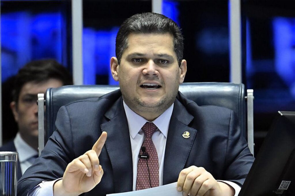 Um homem de terno sentado em cadeira de couro aponta o dedo enquanto fala em reunião