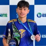 Jovem segura troféu e medalha em frente a painel da Secretaria Municipal de Esporte