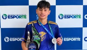 Jovem segura troféu e medalha em frente a painel da Secretaria Municipal de Esporte
