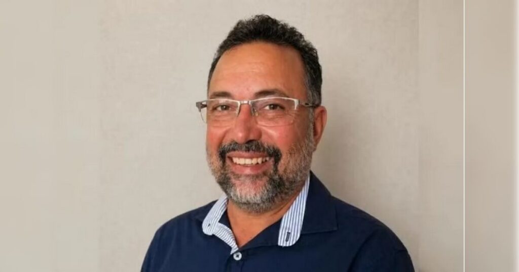 Um homem de óculos e camisa azul sorrindo em frente a parede clara