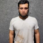 Um homem de camiseta branca com mancha escura em pé diante de parede cinza