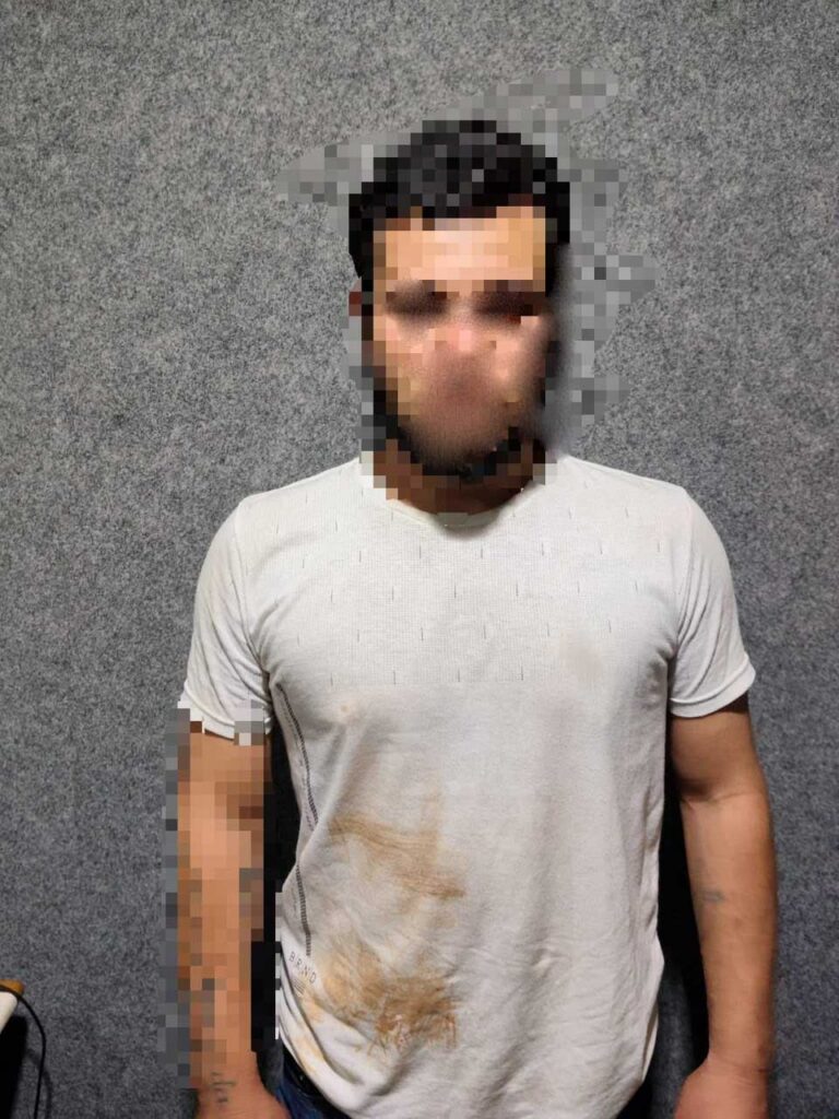 Um homem de camiseta branca com mancha escura em pé diante de parede cinza