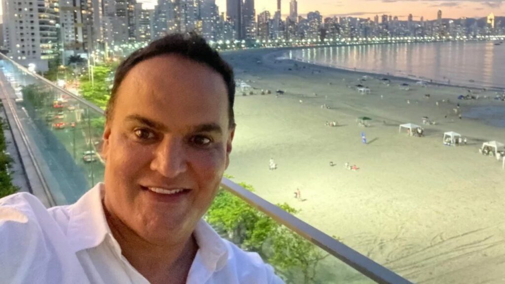 Um homem de camisa branca posa em frente a uma praia com cidade ao fundo ao entardecer