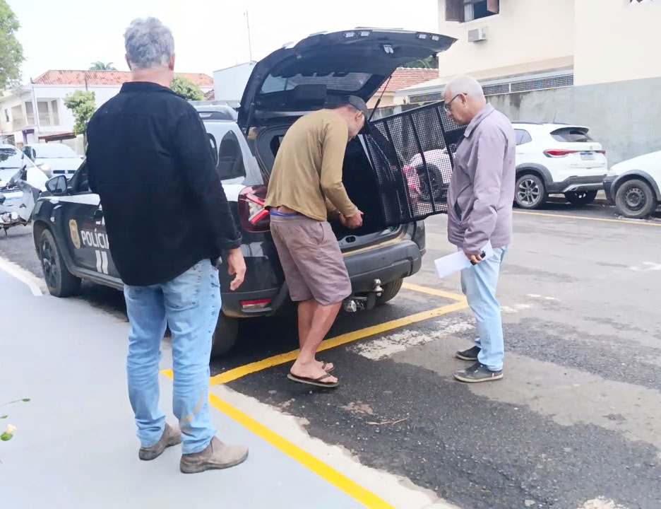 Três homens em frente a um veículo da Polícia com o porta-malas aberto em estacionamento