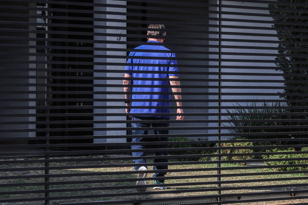 Um homem de camisa azul caminha em frente a uma grade metálica em frente a um prédio