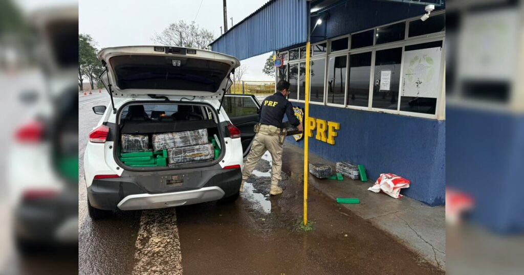 Um homem de uniforme está ao lado de um carro com o porta-malas aberto, revelando pacotes embalados em um local molhado perto de um prédio com letras amarelas na parede