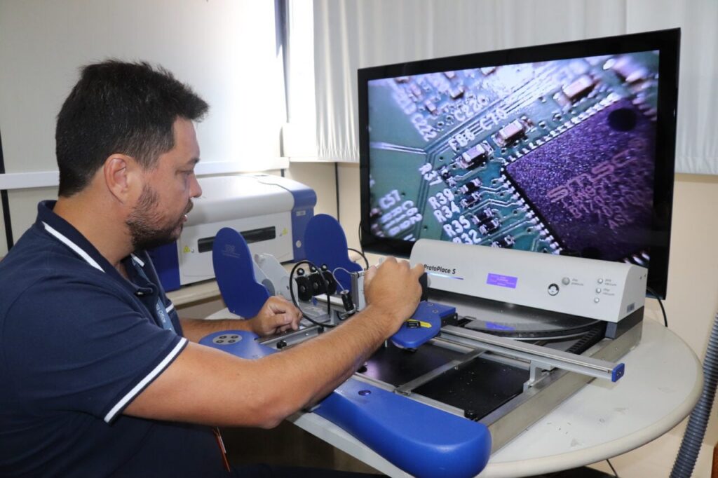 Um homem opera uma máquina de inspeção em um laboratório com uma tela mostrando um circuito eletrônico
