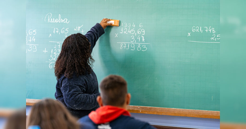 Uma mulher apaga cálculos em uma lousa verde enquanto alunos observam em sala de aula