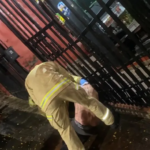 Um bombeiro socorre uma pessoa ferida no chão próximo a um portão metálico em ambiente externo noturno