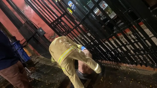 Um bombeiro socorre uma pessoa ferida no chão próximo a um portão metálico em ambiente externo noturno