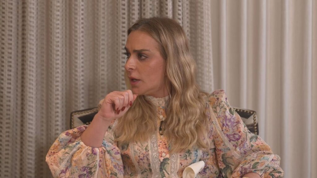 Uma mulher de cabelos loiros e vestido estampado sentada em uma cadeira com as mãos juntas perto do rosto em um ambiente interno com cortinas claras ao fundo