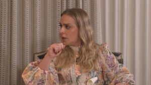 Uma mulher de cabelos loiros e vestido estampado sentada em uma cadeira com as mãos juntas perto do rosto em um ambiente interno com cortinas claras ao fundo