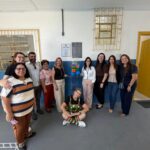 Um grupo de pessoas posa em frente a uma parede com placas e um globo de balões coloridos dentro de uma sala com piso cinza e janelas com grades
