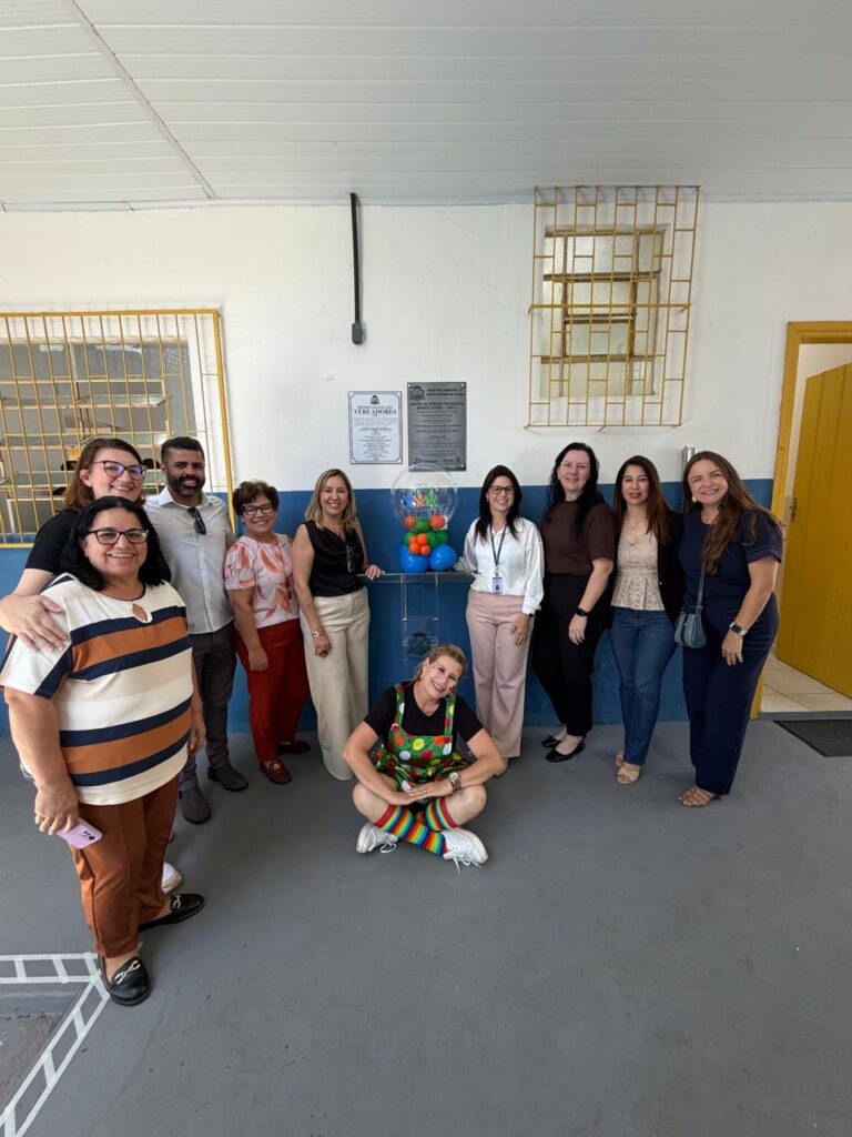 Um grupo de pessoas posa em frente a uma parede com placas e um globo de balões coloridos dentro de uma sala com piso cinza e janelas com grades