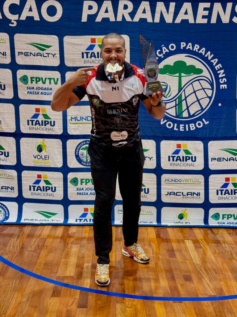 Um homem segura troféu e medalha em quadra de vôlei com banner da Federação Paranaense de Vôlei ao fundo