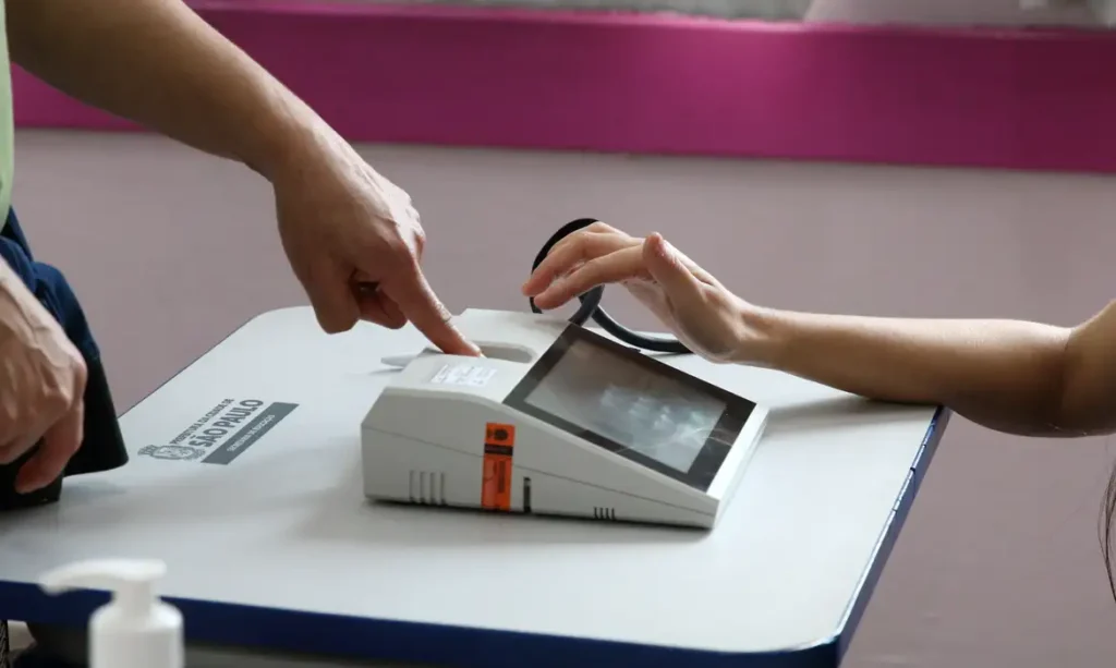 Um homem de terno aponta para uma máquina de votação enquanto uma pessoa posiciona o dedo no leitor biométrico