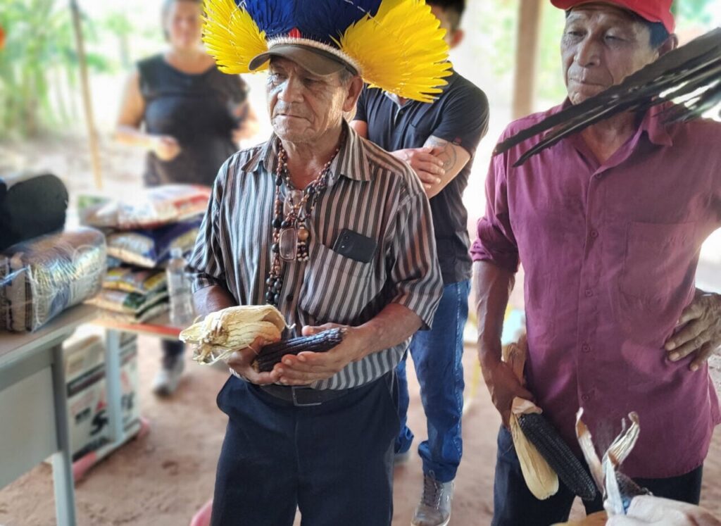 Um grupo de pessoas segura espigas de milho em um ambiente coberto com sacos de sementes ao fundo