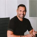 Um homem de camiseta preta sentado em uma cadeira em um ambiente interno