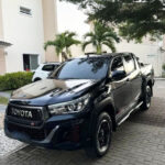 Camionete preta da marca Toyota estacionada em pátio com calçamento de pedra e vegetação ao fundo