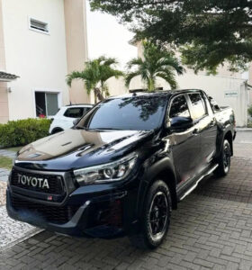 Camionete preta da marca Toyota estacionada em pátio com calçamento de pedra e vegetação ao fundo