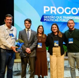 Cinco pessoas posam em palco com troféu e crachás, ao fundo painel com texto "PROGOV Gestão que transforma políticas públicas