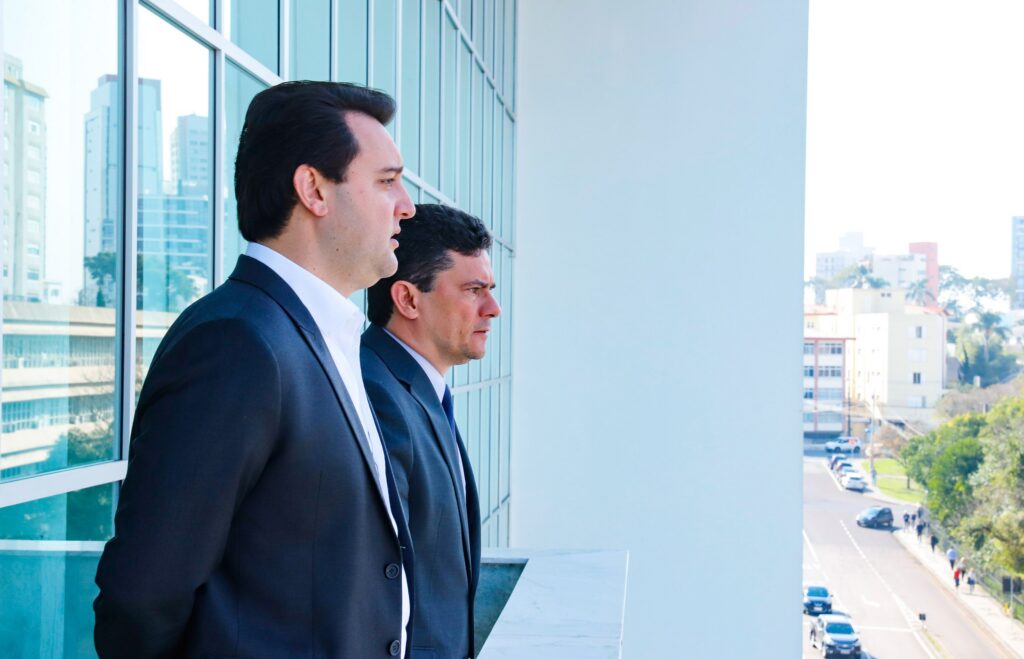 Dois homens de terno observam uma via urbana de um terraço com vista para edifícios