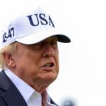 Um homem de terno e camisa branca usa boné branco com as letras USA e números 45-47